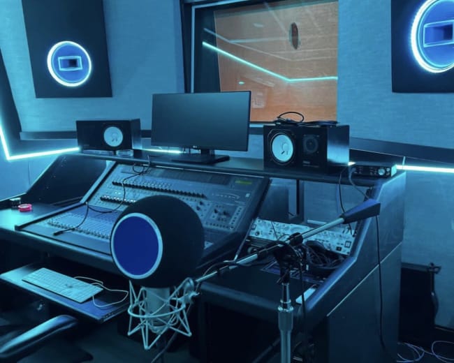 The image shows a modern recording studio with a mixing console, monitor screens, and a microphone setup, illuminated by blue LED lighting.