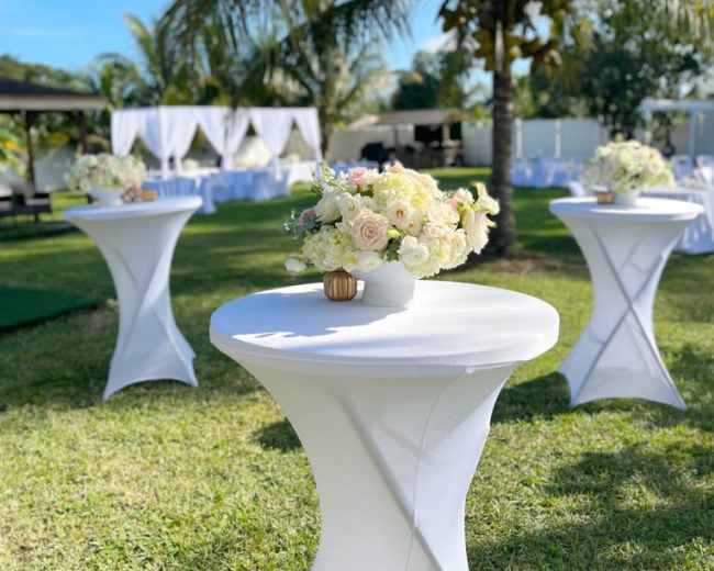 The scene features several white cocktail tables adorned with floral centerpieces set on a grassy area surrounded by palm trees and a clear blue sky.