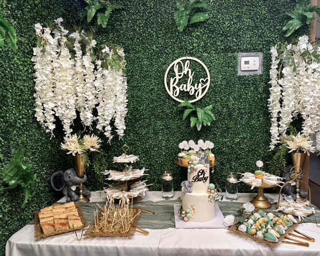 A decorated table displays a tiered cake, cookies, and snacks, surrounded by greenery and floral arrangements for a baby celebration.