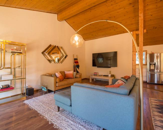 The living room features a beige sofa, a light blue sectional, a gold shelving unit, and a large wall-mounted television, all under a wooden ceiling.