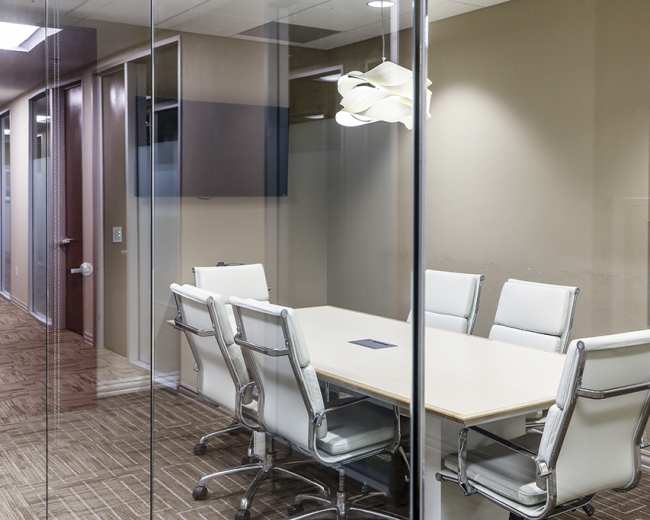 A conference room with a glass wall features a long table surrounded by six white leather chairs.