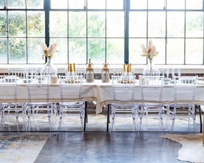 A long dining table is set with glassware and floral centerpieces, positioned in front of large windows that allow natural light to fill the space.