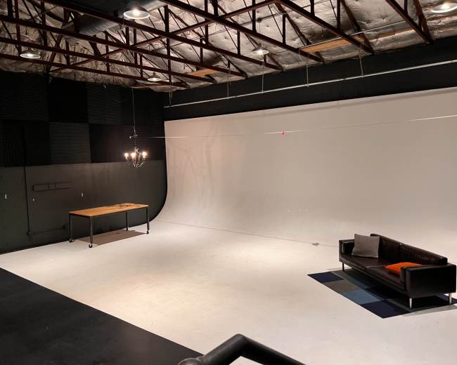 The image shows a spacious photography studio with a white backdrop, a wooden table, and a black couch against the wall.