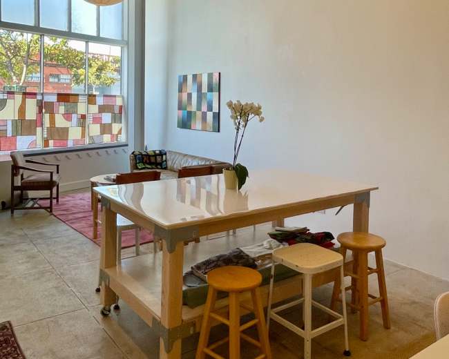 The image shows a spacious, well-lit room with a large white table, wooden stools, and decorative window panes.