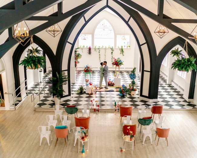 A couple embraces on a decorated stage in a spacious indoor venue with black and white checkered flooring and floral arrangements.