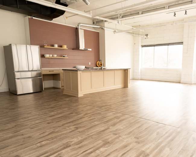 The image shows an empty kitchen space with a large central island, a stainless steel refrigerator, and light-colored wood flooring.
