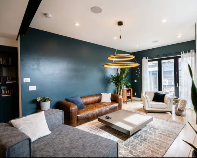A modern living room features a brown leather sofa, a gray sectional, a round coffee table, and large windows with drapes, complemented by greenery and contemporary lighting fixtures.