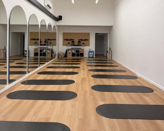 The image shows a spacious yoga studio with wooden floors, several yoga mats arranged in parallel, and large mirrors reflecting the room.