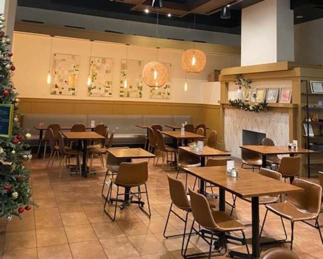 The image shows an empty café with wooden tables and chairs, illuminated by pendant lights and featuring a decorative Christmas tree in the corner.