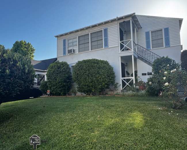 A two-story house with a light blue exterior and a front staircase is surrounded by well-maintained greenery and lawn.
