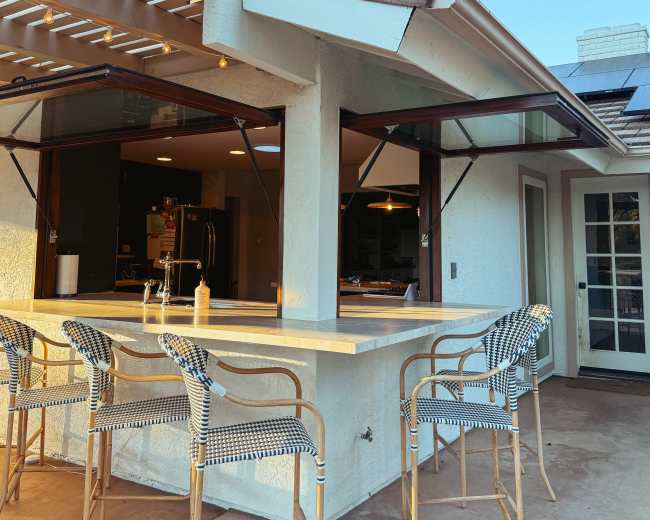 The image shows a modern outdoor bar area with several wicker chairs, a countertop, and large windows that open to an indoor kitchen space.