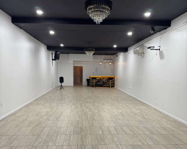 The image shows an empty hall with tiled flooring, white walls, and a black ceiling, featuring a small bar area with lighting at the far end.