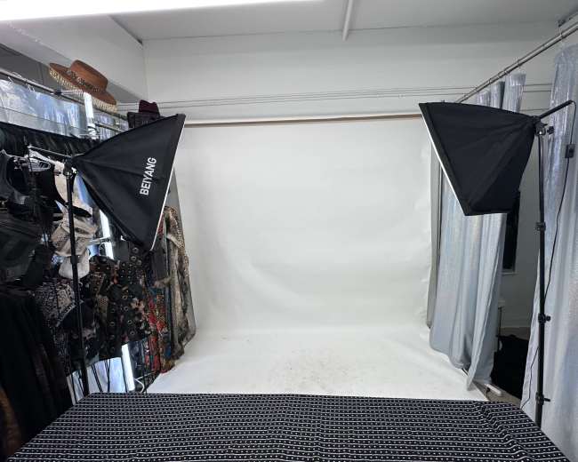 The image shows a photography setup with two softbox lights flanking a backdrop of white paper, surrounded by clothing racks on either side.
