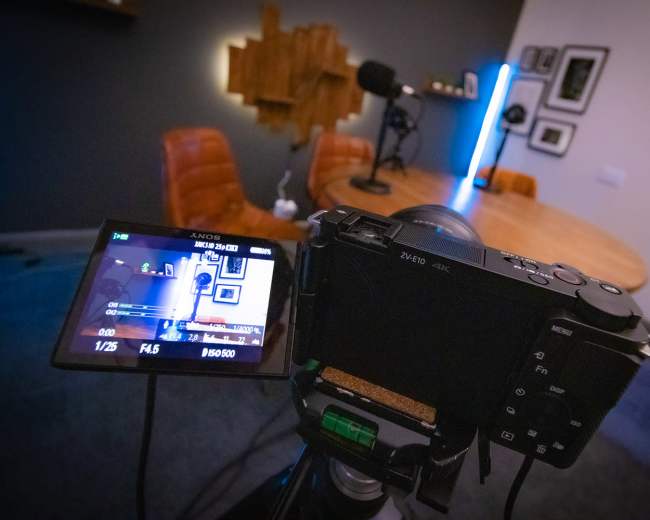 A camera is positioned on a tripod, facing a table surrounded by chairs in a room set up for video recording.