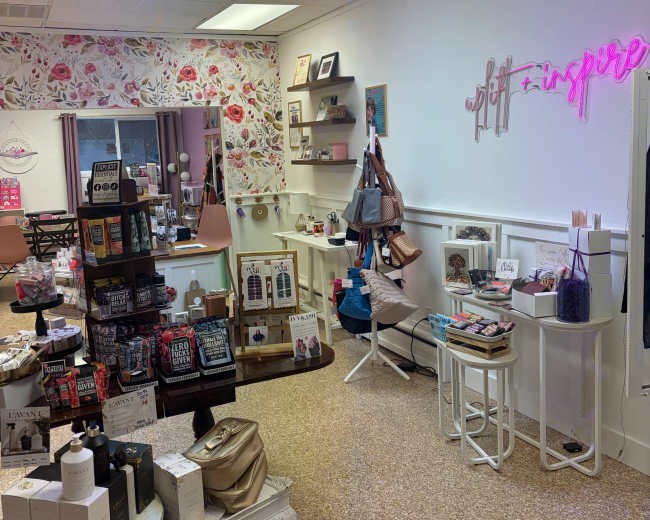 The image shows a boutique shop filled with various gift items, decor, and displays, featuring floral wallpaper and a neon sign that reads "gift + inspire."