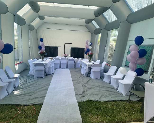 A large, inflatable tent is set up with tables and chairs arranged for an event, decorated with balloons and flowers.