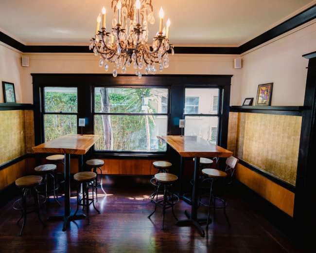 The image shows a cozy room with wooden flooring, a chandelier, and seating at tall tables near large windows.