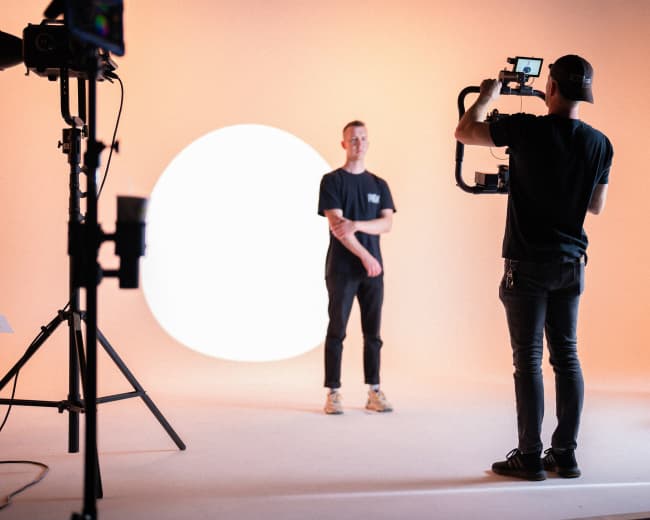 A man stands in front of a circular light while another man with a camera captures the scene in a studio setting.
