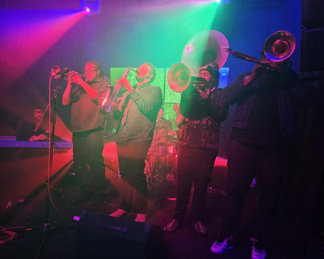 A group of musicians plays brass instruments on stage, illuminated by colorful stage lights.