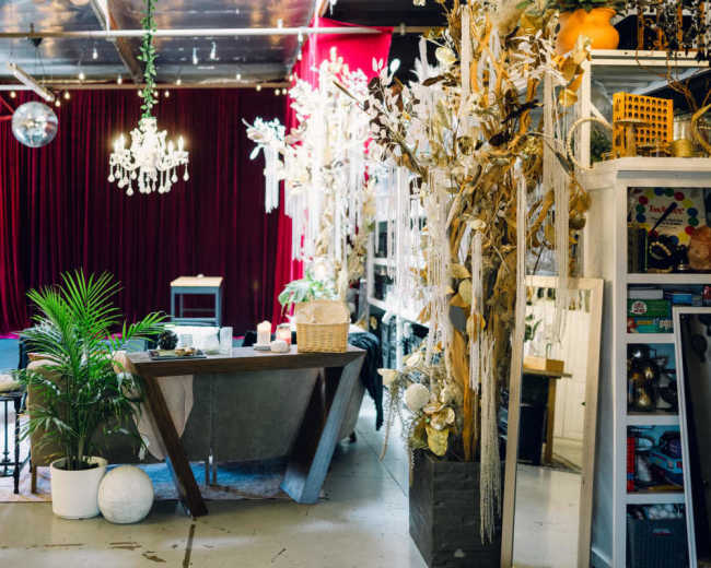 The image depicts a spacious event venue featuring a table at the center, surrounded by decorative plants, with a red velvet backdrop and various artistic installations along the walls.
