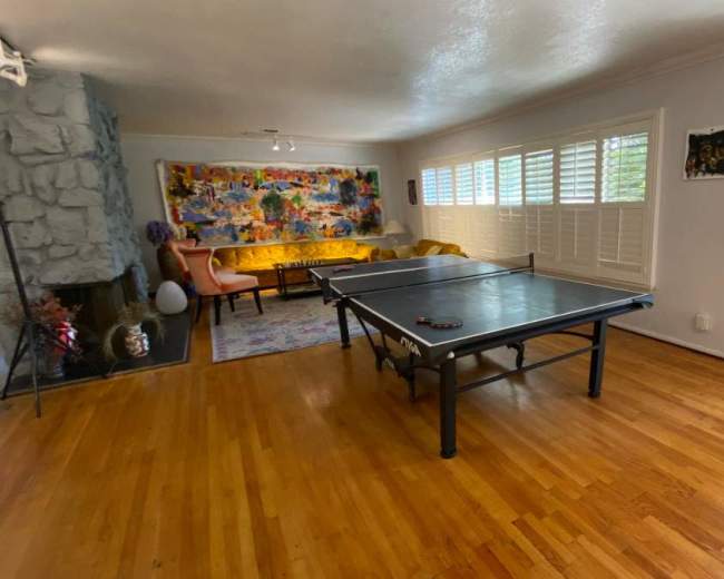 The image shows a spacious living room featuring a ping pong table, hardwood flooring, and a colorful artwork on the wall.