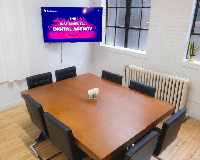 A conference room features a large wooden table surrounded by black chairs, with a screen on the wall displaying "THE INSTRUMENTAL DIGITAL AGENCY."