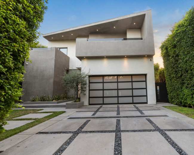 A modern two-story house features a sleek design with a garage and a driveway lined by gravel, surrounded by tall green hedges.