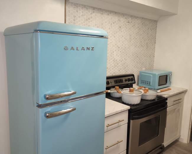 The image shows a kitchen featuring a retro-style light blue refrigerator beside an oven and a matching microwave, with a tiled backsplash and white cabinetry.