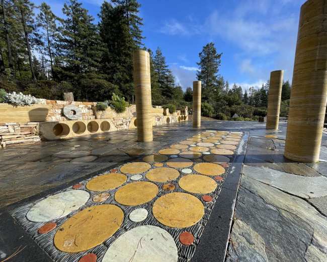 The image shows a pathway with circular stone designs and several tall columns surrounded by trees and a blue sky.