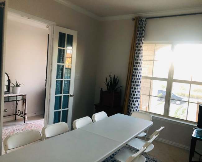 A bright room features a long white table surrounded by chairs, with a door leading to another room and a window showing a glimpse of a car outside.