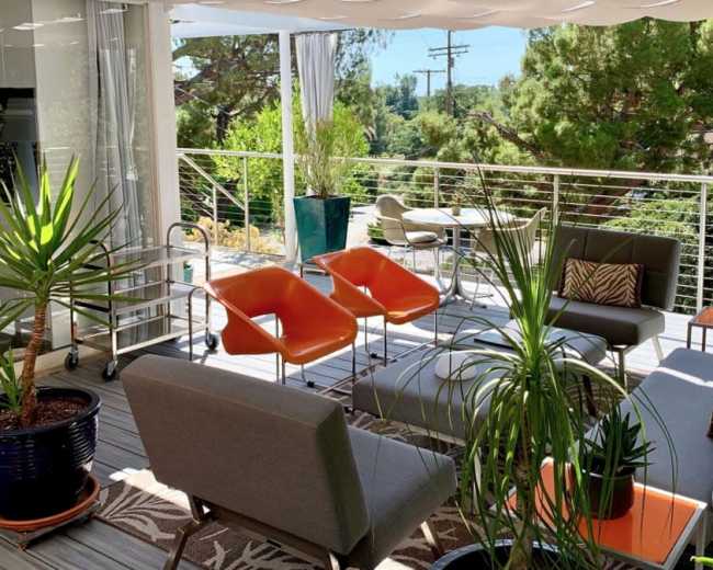 The image shows a modern outdoor patio with sleek furniture, potted plants, and a view of greenery in the background.