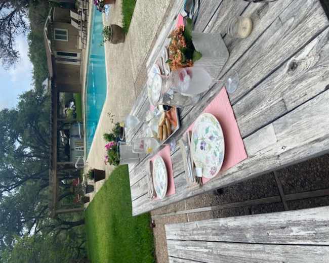 A rustic wooden table is set with plates and drinks beside a clear swimming pool in a garden area.
