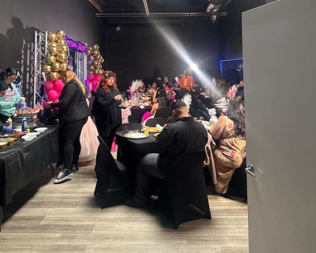 The image shows a lively indoor event with guests seated at tables, enjoying food and celebrating, surrounded by colorful decorations and balloons.