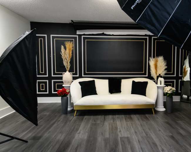 A well-lit photography studio featuring a white sofa with black pillows, flanked by large softbox lights and decorative vases with plants.