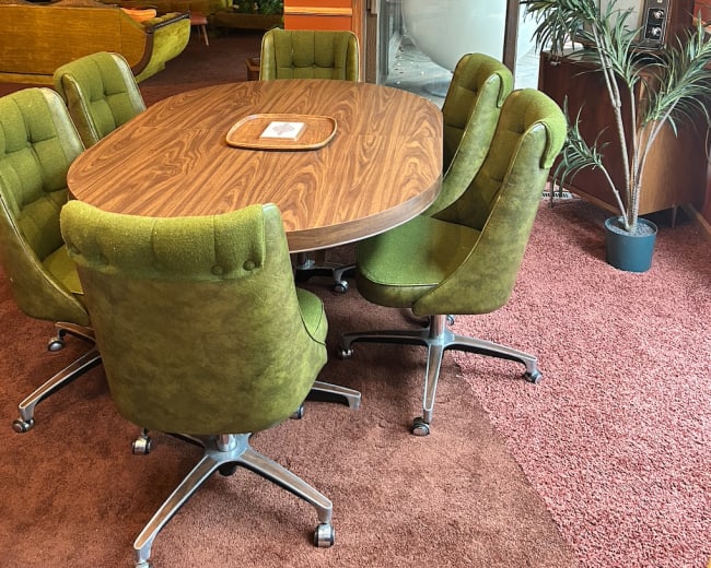 The image shows a round wooden dining table surrounded by six green upholstered chairs in a room with reddish-brown walls and ambient lighting.