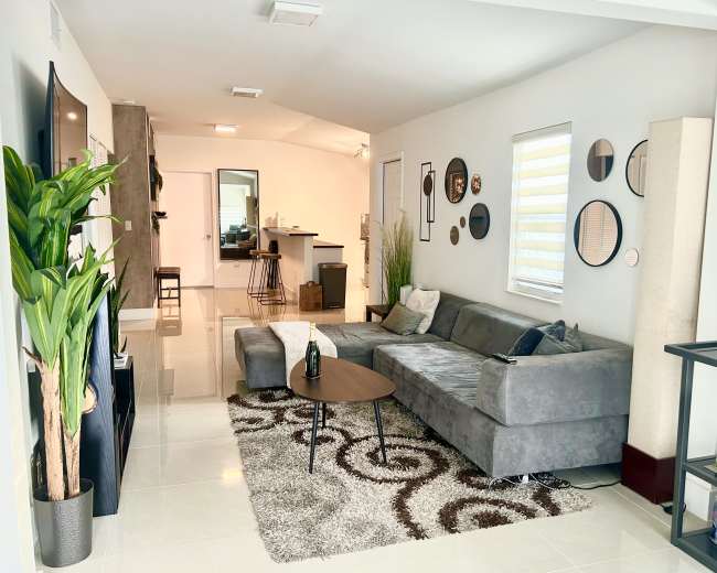 A modern living room featuring a gray sectional sofa, a round coffee table, and decorative mirrors on the walls, with a small kitchen area visible in the background.