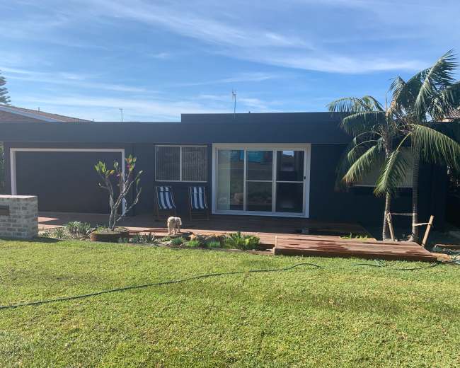 A modern black house with a wooden deck, large windows, and palm trees is set against a green lawn.
