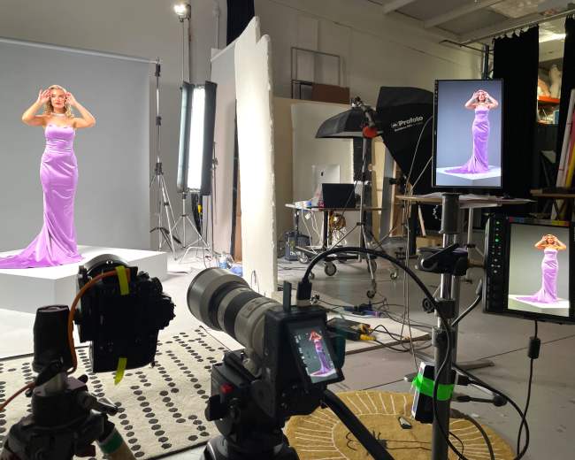 A model poses in a lavender gown on a pedestal in a photography studio, surrounded by lighting equipment and cameras.