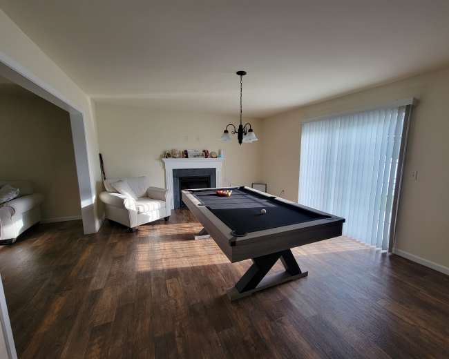 A spacious room featuring a billiard table, a fireplace, and two armchairs near a window with vertical blinds.