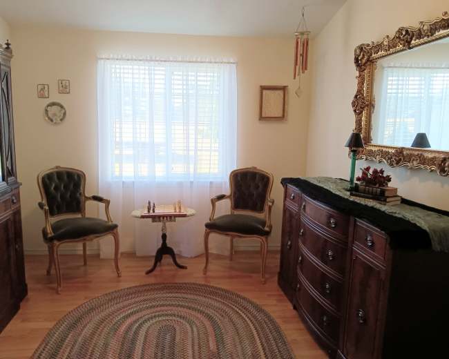 The image shows a cozy room with two upholstered chairs, a small table between them, a decorative mirror on the wall, and a dresser adorned with a plant.