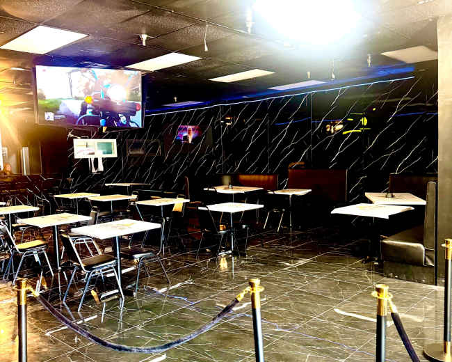 The image shows an empty indoor dining area with tables and chairs arranged around a black and white marble floor, and a television mounted on a wall displaying a game.