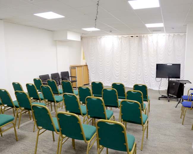 The image shows a sparsely furnished room with rows of green chairs facing a television and a curtain backdrop.