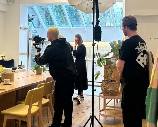 A camera crew is set up in a bright café, with one person filming while another adjusts lighting equipment as a third person observes.