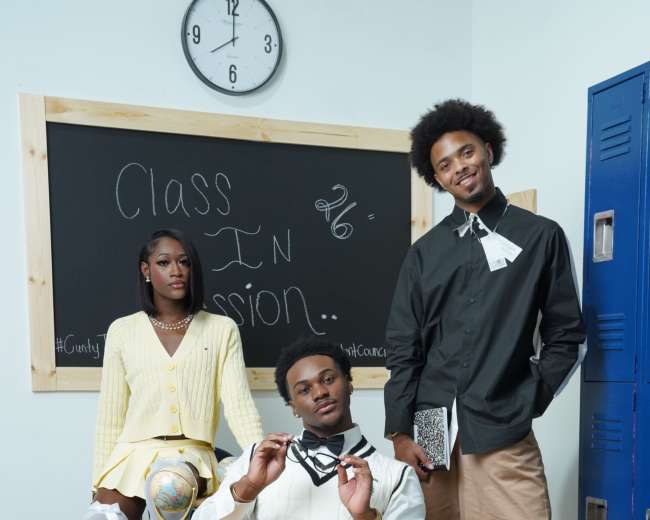 Three young adults pose in a classroom setting, with one seated at a desk and the others standing beside it, dressed in stylish outfits reminiscent of a school theme.