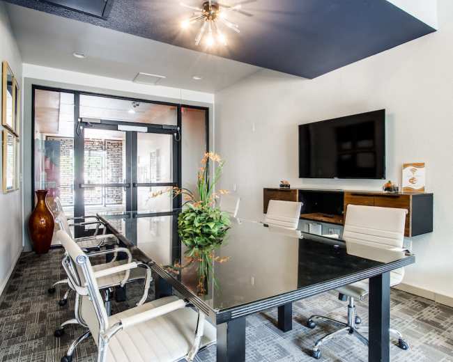 A modern conference room features a glass table surrounded by white chairs, with a television mounted on the wall and large windows allowing natural light.