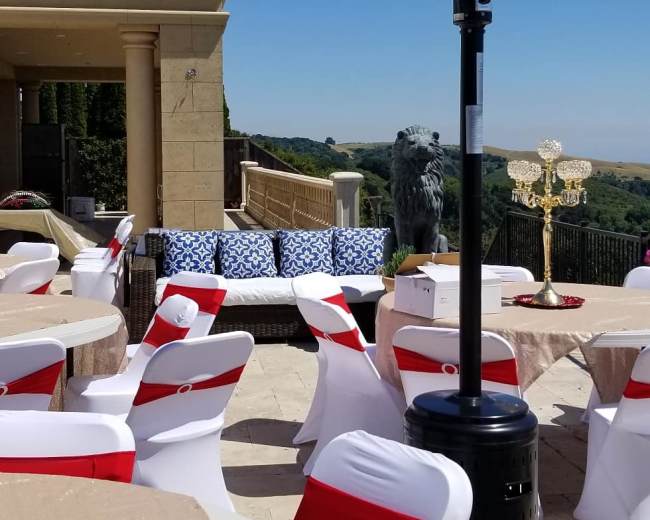 The patio features round tables covered with beige tablecloths, white chairs with red sashes, and a blue and white patterned sofa, with a decorative candelabra and a lion statue in the background.