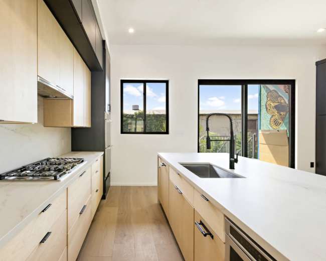 The image shows a modern kitchen with wooden cabinets, a gray countertop, and windows providing natural light.