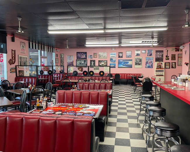 The interior of a retro-style diner features red booths, black and white checkered flooring, and walls adorned with vintage posters and records.