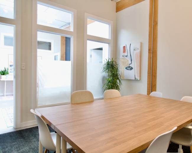 The image shows a modern conference room with a large wooden table and white chairs, featuring a plant and artwork on the wall.