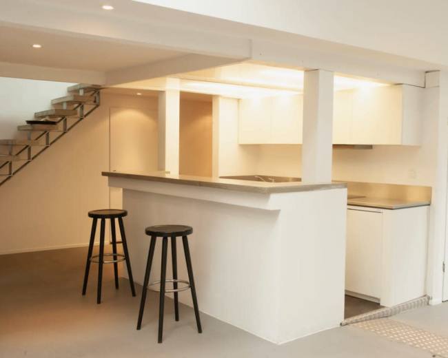 The image shows a modern kitchen with a minimalist design, featuring a bar area with two black stools and a staircase in the background.